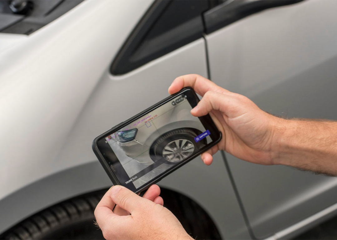 Two hands holding a smart phone aimed at a silver car, the phone's screen shows an AI-powered scan of possible vehicle damage.