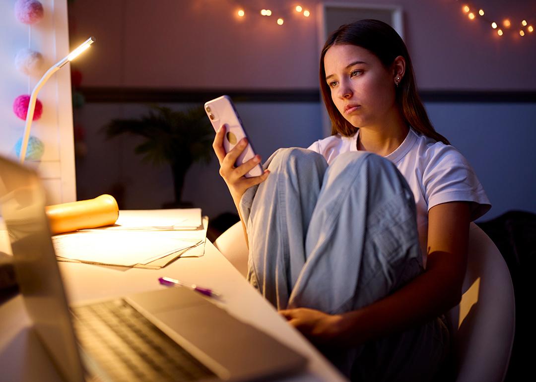 A young woman sitting at a desk in her bedroom and browsing her phone at night.