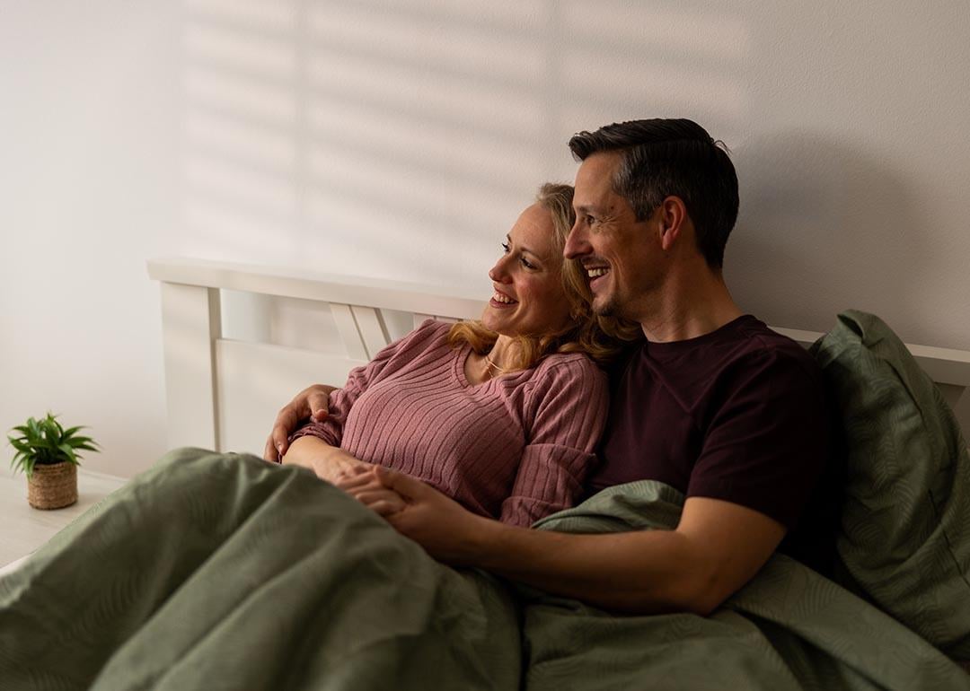 A couple sitting in bed wrapped in blankets.