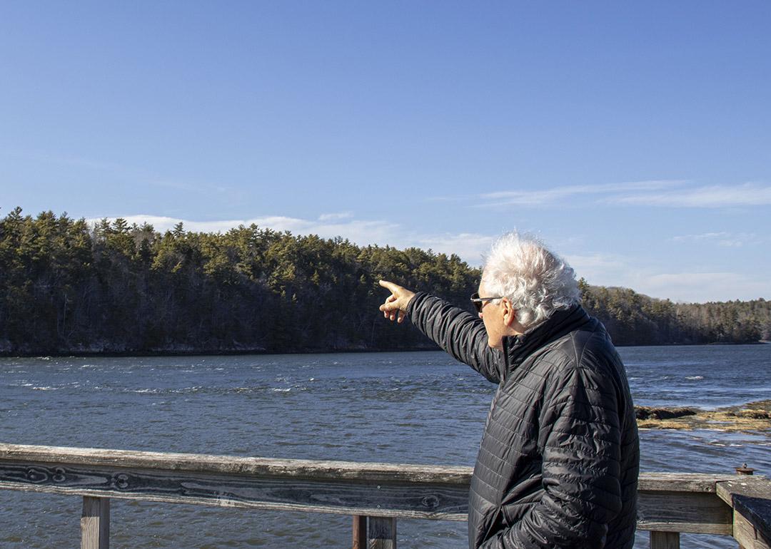 Protect Wiscasset co-founder Richard Davis facing away from the camera wearing a black jacket and his white air blown by the wind, points to the undeveloped land across a waterway where the prospect of a $5 billion data center emerged.