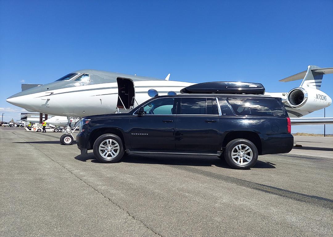 A black Chevrolet Suburban car parked alongside a private air jet in an open ground.