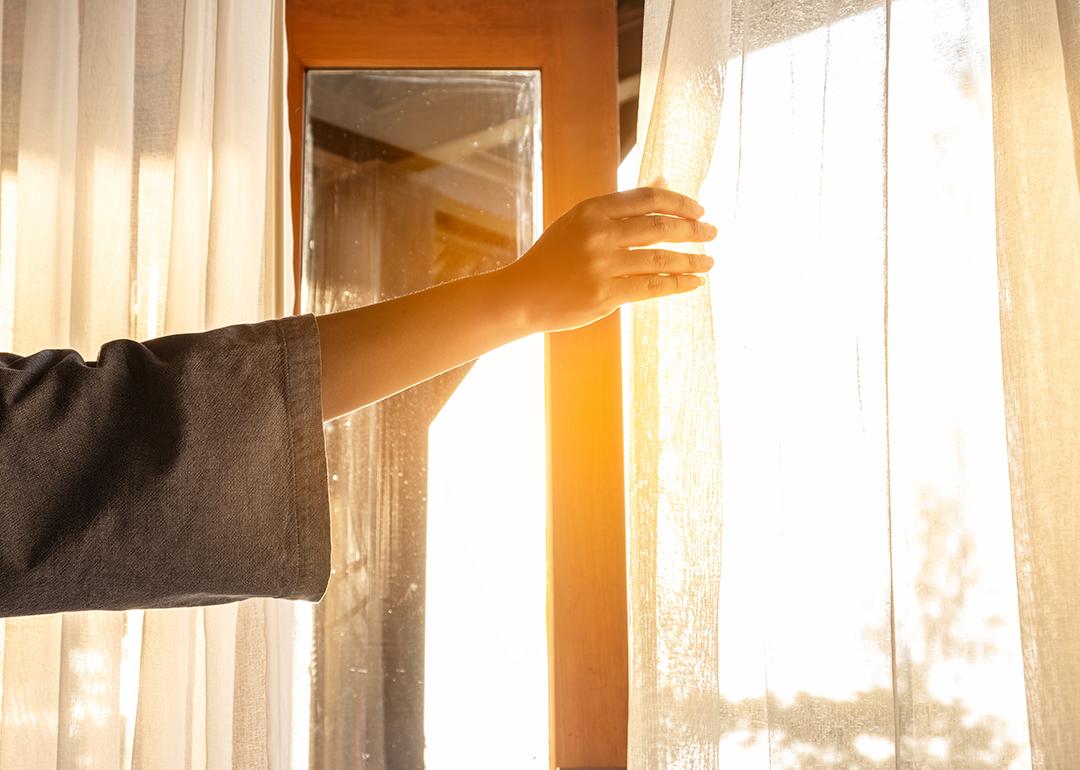 A woman's hand opening the curtains to a window to let in sunlight.