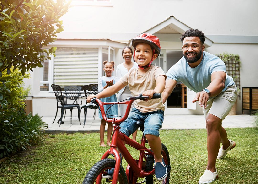 A young boy learning how to ride a bicycle being cheered on by his family.