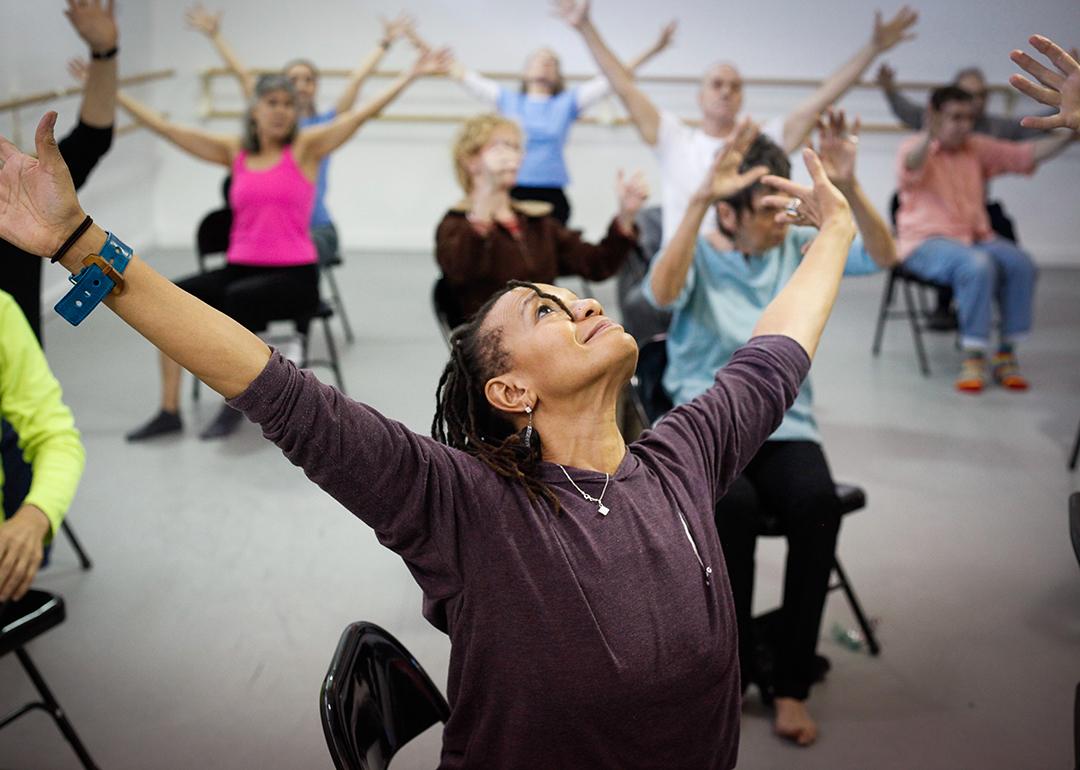 View of upturned face of Black woman with her arms extended, leading a chair dance class with lots of seniors sitting in the background extending their arms in the same motion.