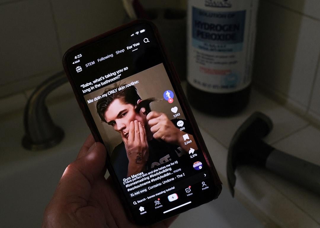A person watching from his smartphone a video of a male TikTok influencer hammering his cheekbone, while a hydrogen peroxide bottle and a hammer are also seen in his bathroom.