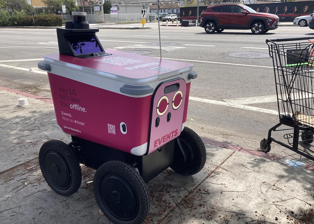 A pink sidewalk delivery robot built by California-based Serve Robotics features an ad from dating app Tinder, while sitting on a sidewalk next to a shopping cart with cars on the road in the background.