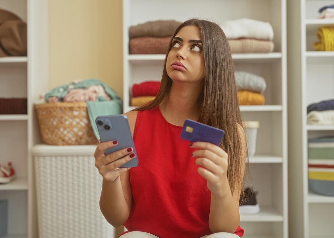 A young adult appearing to be frustrated while holding her smartphone and credit card in a dressing room.