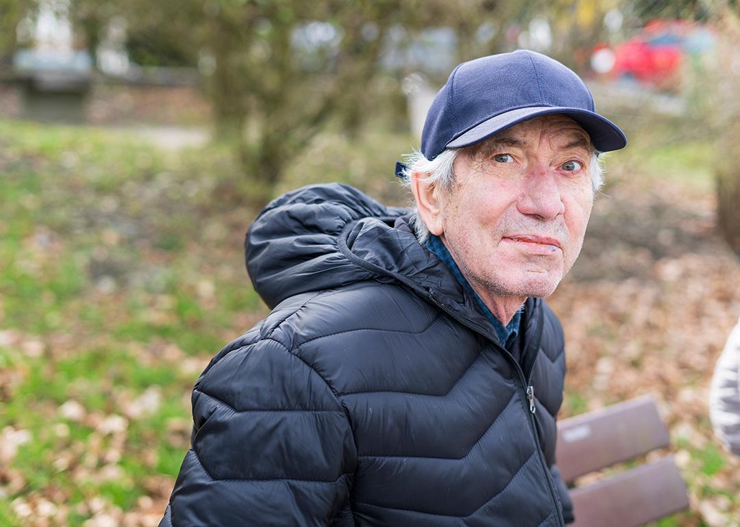 An elderly man wearing a thick jacket and hat while sitting alone on a bench in a park.