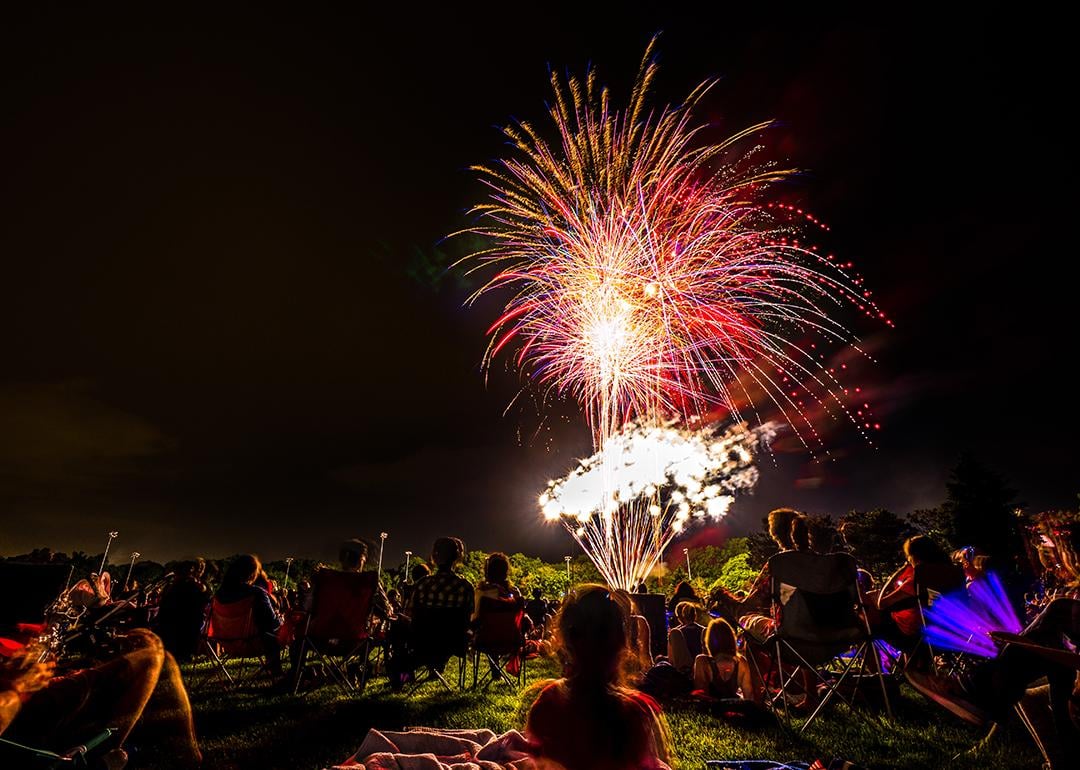Crowds of people sit in lawn chairs and blankets to enjoy the colorful fourth of July independence day celebratory fireworks exploding in the black night sky.