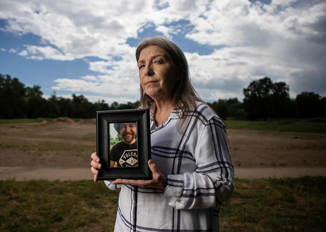 Jeanelle Couch holds framed photo of her son, David Couch, who was killed in a shooting involving a California Highway Patrol officer. She is standing in Cascade Park in Redding with a sky full of clouds and open space behind her.