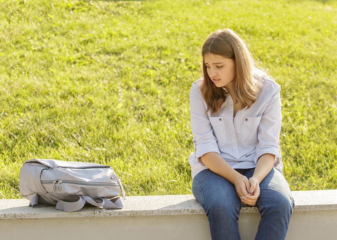 An upset teenage girl sitting on a curb and staring at her backpack for school.