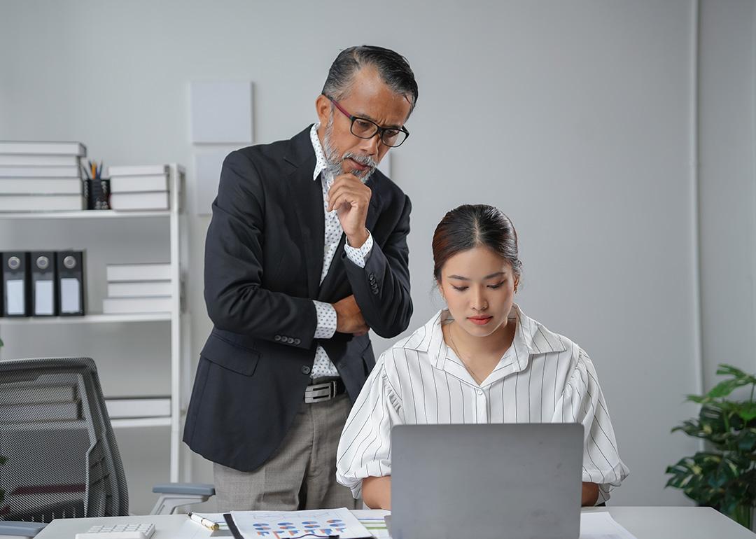 A manager standing behind an employee while checking her work report's progress.