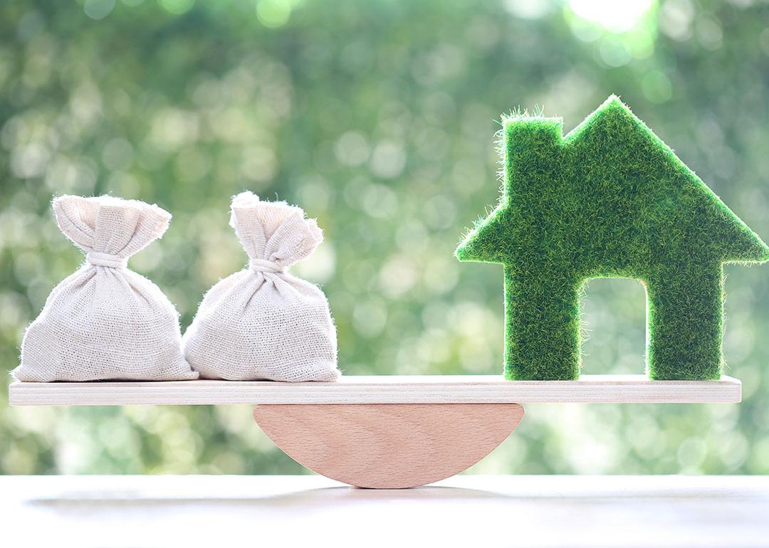 A bag of money and a figure of a home on both ends of a seesaw as a concept of weighing finances and home equity.