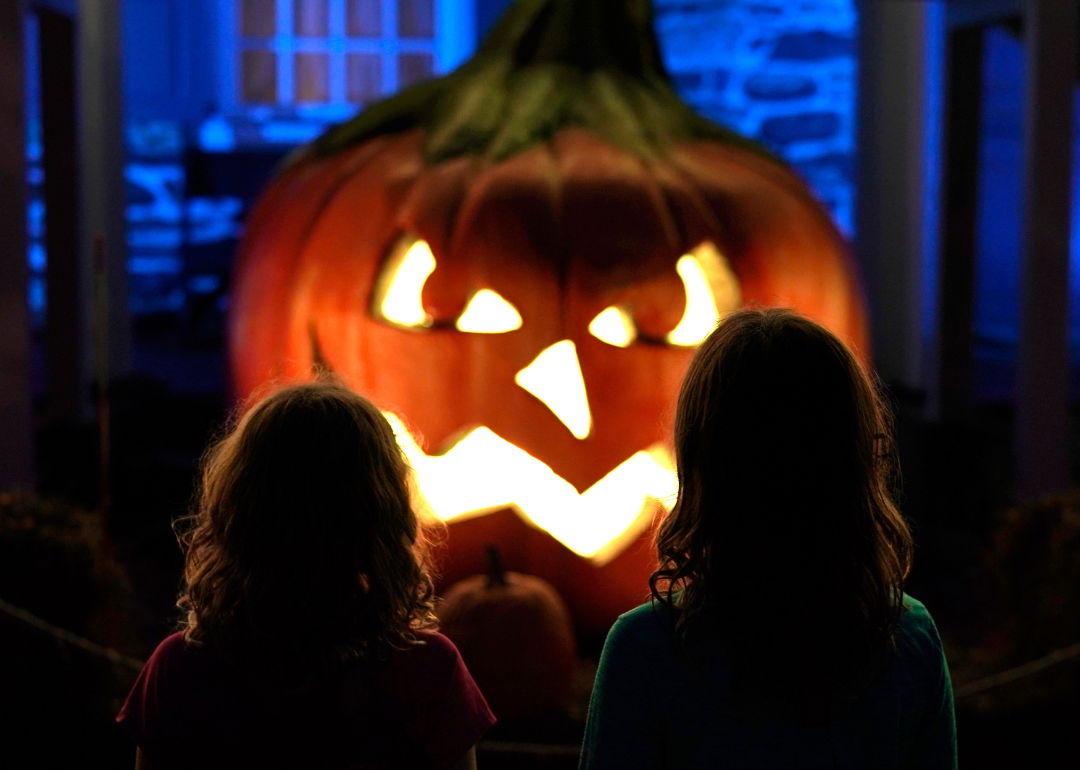 Two young girls standing in front of a giant lit up jack-o'-lantern at night.