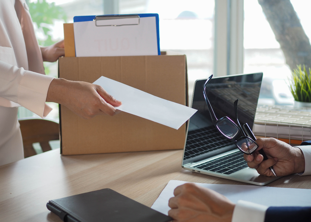 Person puts box of personal belongings on desk and hands boss resignation letter.