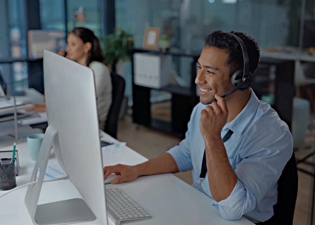 A young man smiling, using a headset while looking at the computer in a modern office.