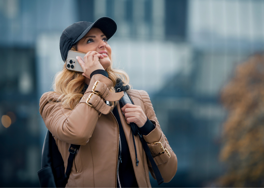 A woman in a black baseball hat and cream jacket holding a cellphone to her ear and looking up. 