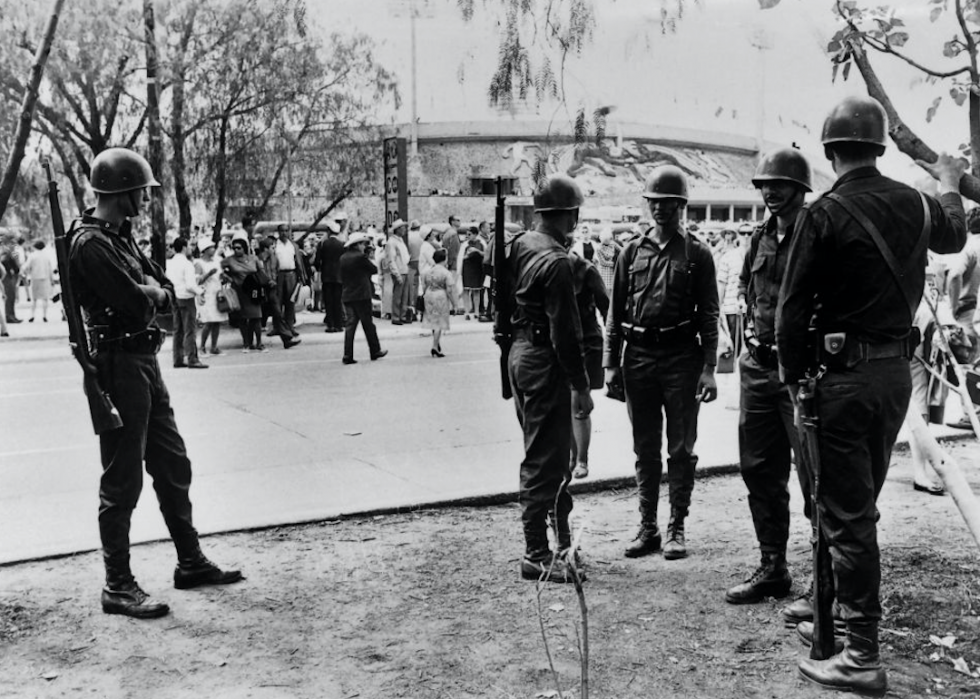 Several men dressed in military outfits stand and talk as protestors are gathered in the background.