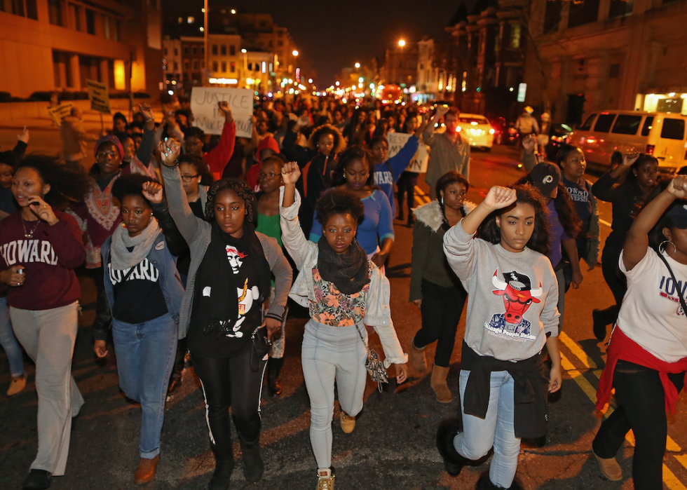 At night, young people walk down the street with their fists in the air. One protestor in the background is holding a sign that says Justice for Mike Brown.