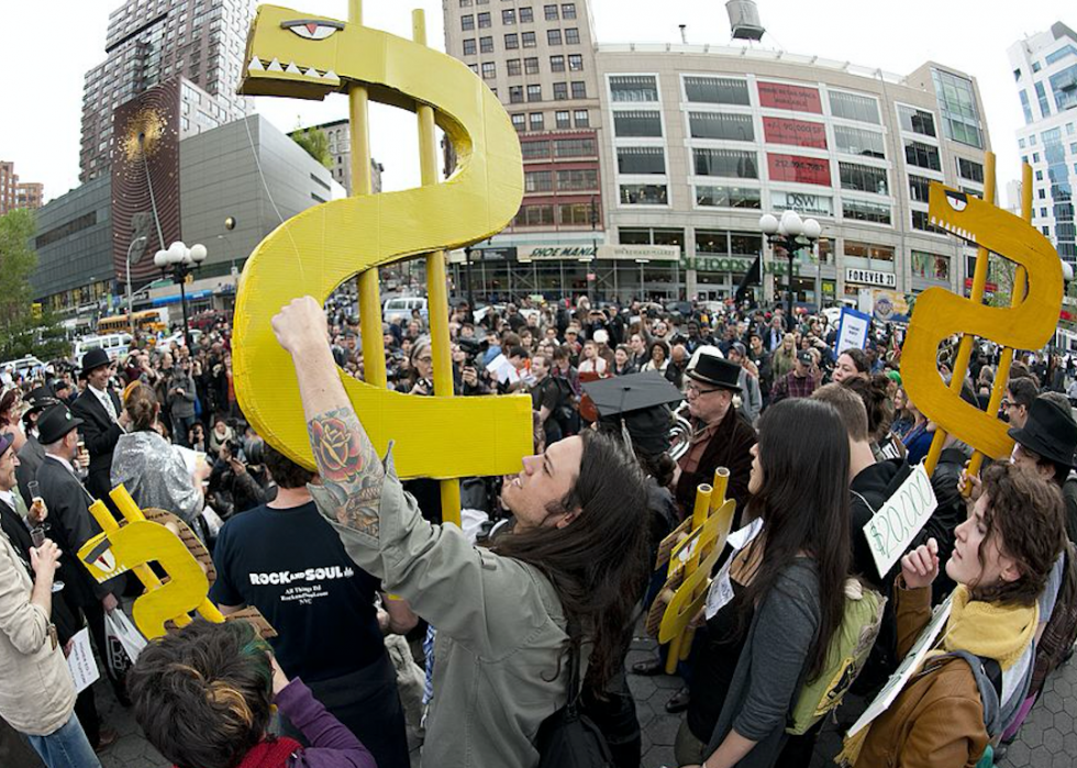 Crowds of protestors stand in the streets with a couple of people in the foreground holding up large, yellow dollar signs with evil eyes and sharp teeth.