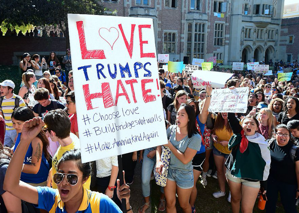 Young people standing in crowds yell, with some holding signs, including one that says Love Trumps Hate! with three hashtags: choose love, build bridges not walls, make America kind again. 