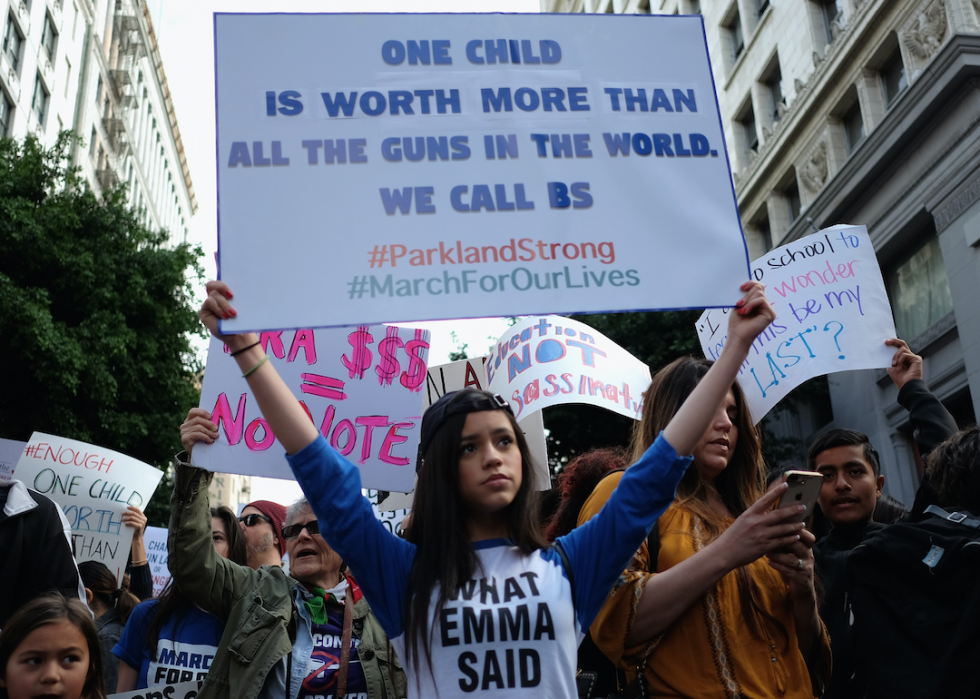 Protestors hold signs, with one young person wearing a shirt that says, what Emma said, holding a sign that says one child is worth more than all the guns in the world, we call B.S., with two hashtags: Parkland Strong and March For Our Lives. 