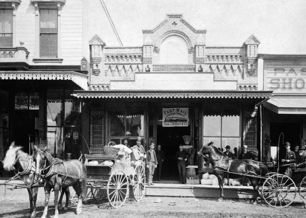 Men standing next to horse drawn carriages in front of shops