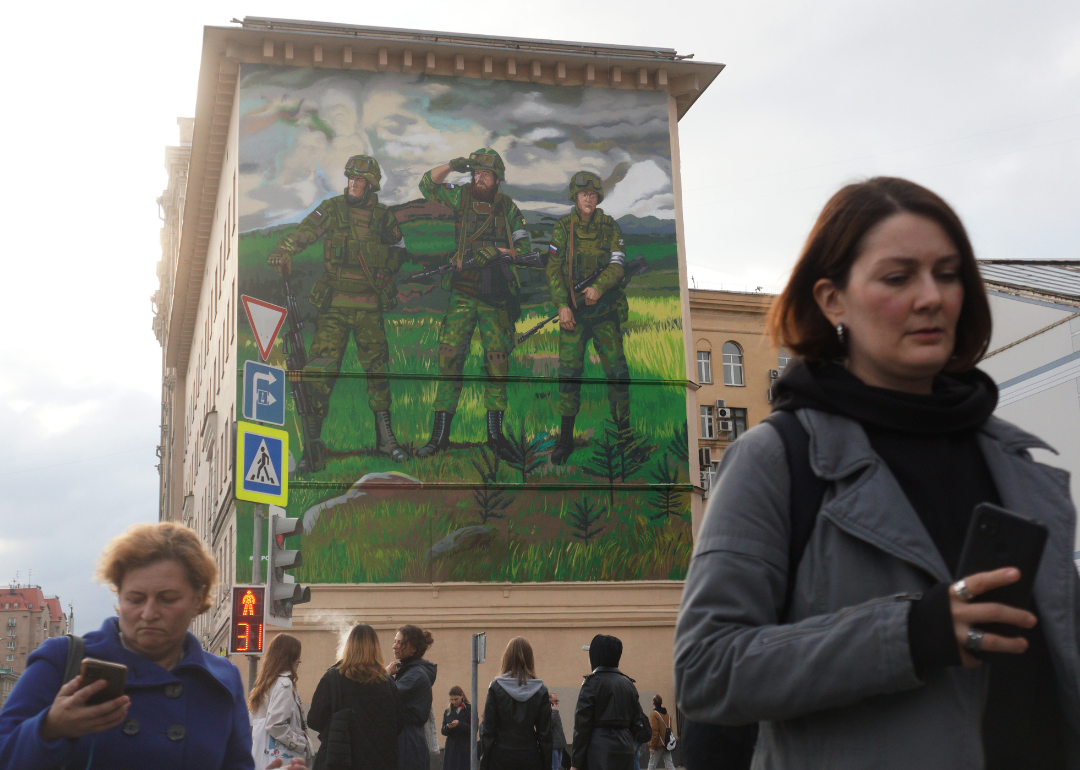 People crossing street in with Moscow with large pro-war mural behind them.