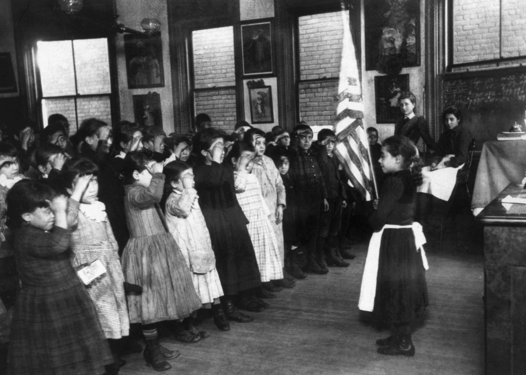 A young girl stands before a classroom of students who salute the American flag.