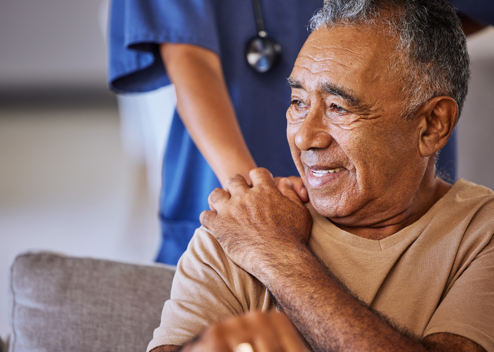 A caregiver standing behind a smiling older man with their hand on his shoulder.