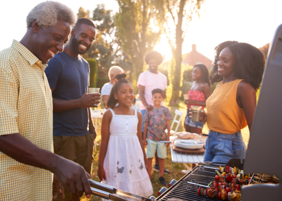 A grandfather figure BBQs with smiling family members gathered around him.