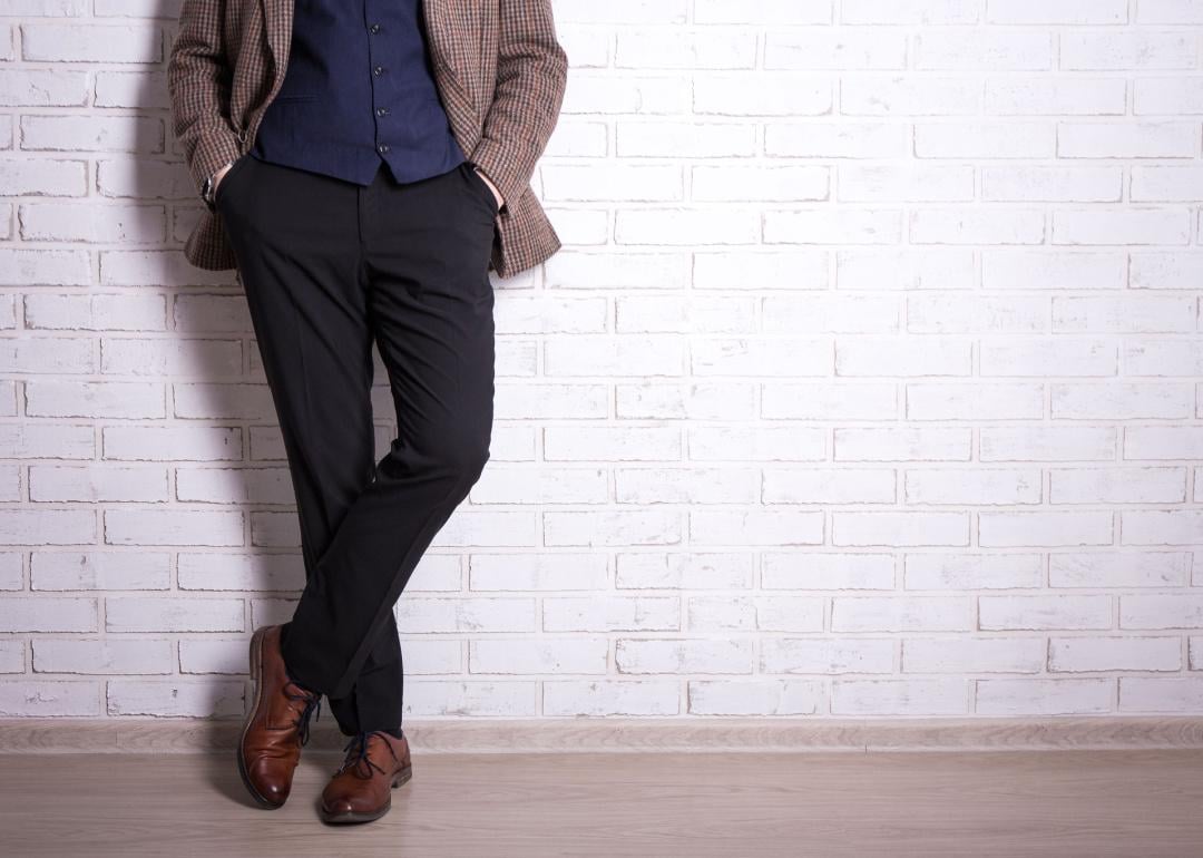 Man dressed in pants, jacket, vest and shoes leaning against wall