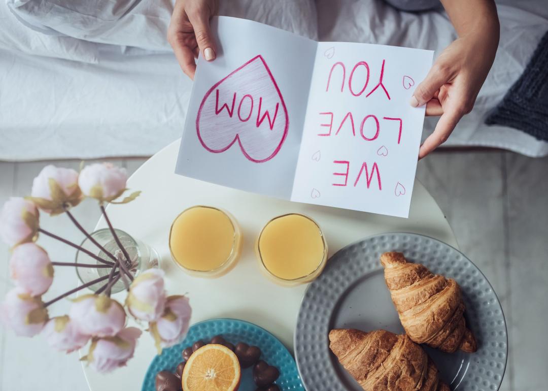 Flowers and breakfast in bed with hands holding ‘We love you mom” card.