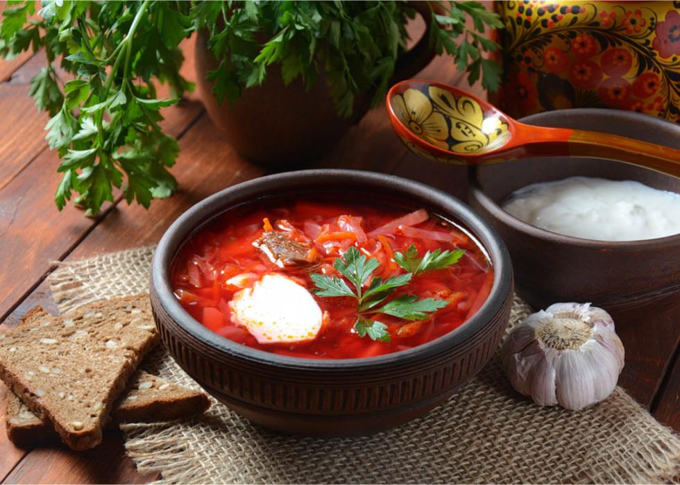 A bowl of borscht with slices of bread and a bowl of sour cream next to it, along with a bulb of garlic and a plant pot of parsley. 