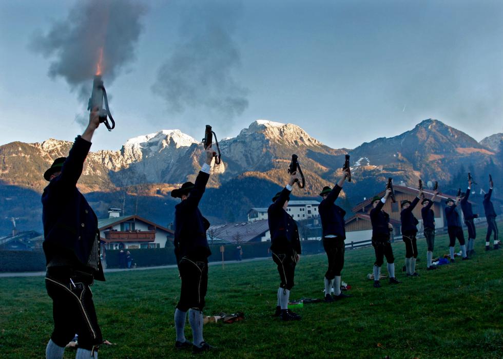Several men in lederhosen firing mortars into the air with a backdrop of houses and snow capped mountains.
