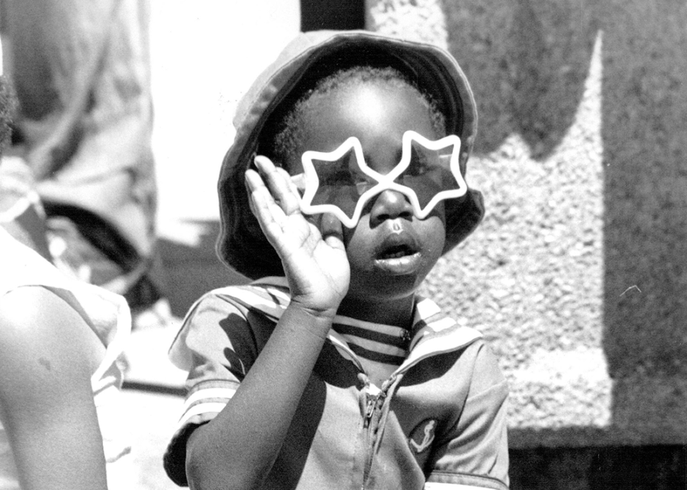 A child wearing star sunglasses waves at a passing parade.