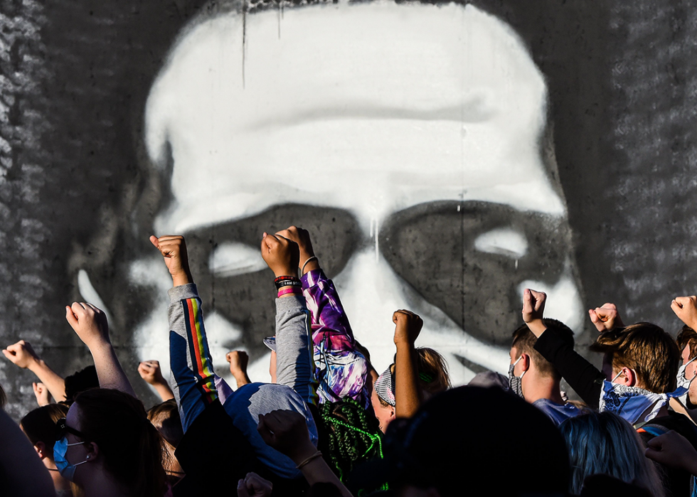 Protestors, some wearing masks, stand with their fists in the air. In the background, partially obscured by people, is a large spray-painted image of a face.