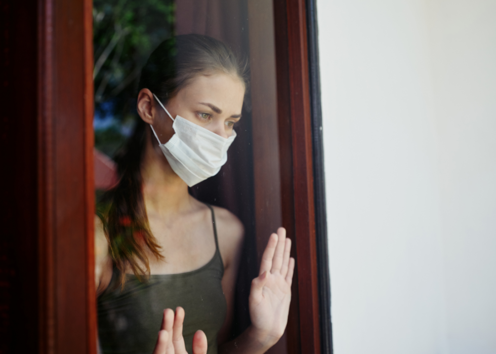 A view from outside a window shows a somber-looking young woman wearing a mask and placing her palms on the window. 