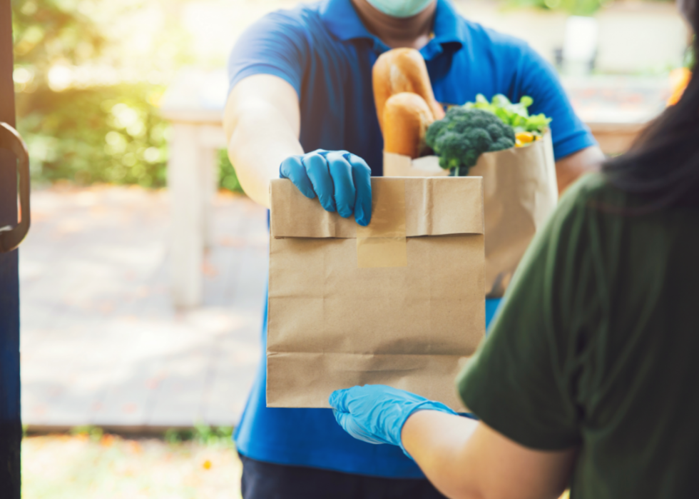 A person accepting groceries from a delivery person. Both people are wearing blue surgical gloves. 