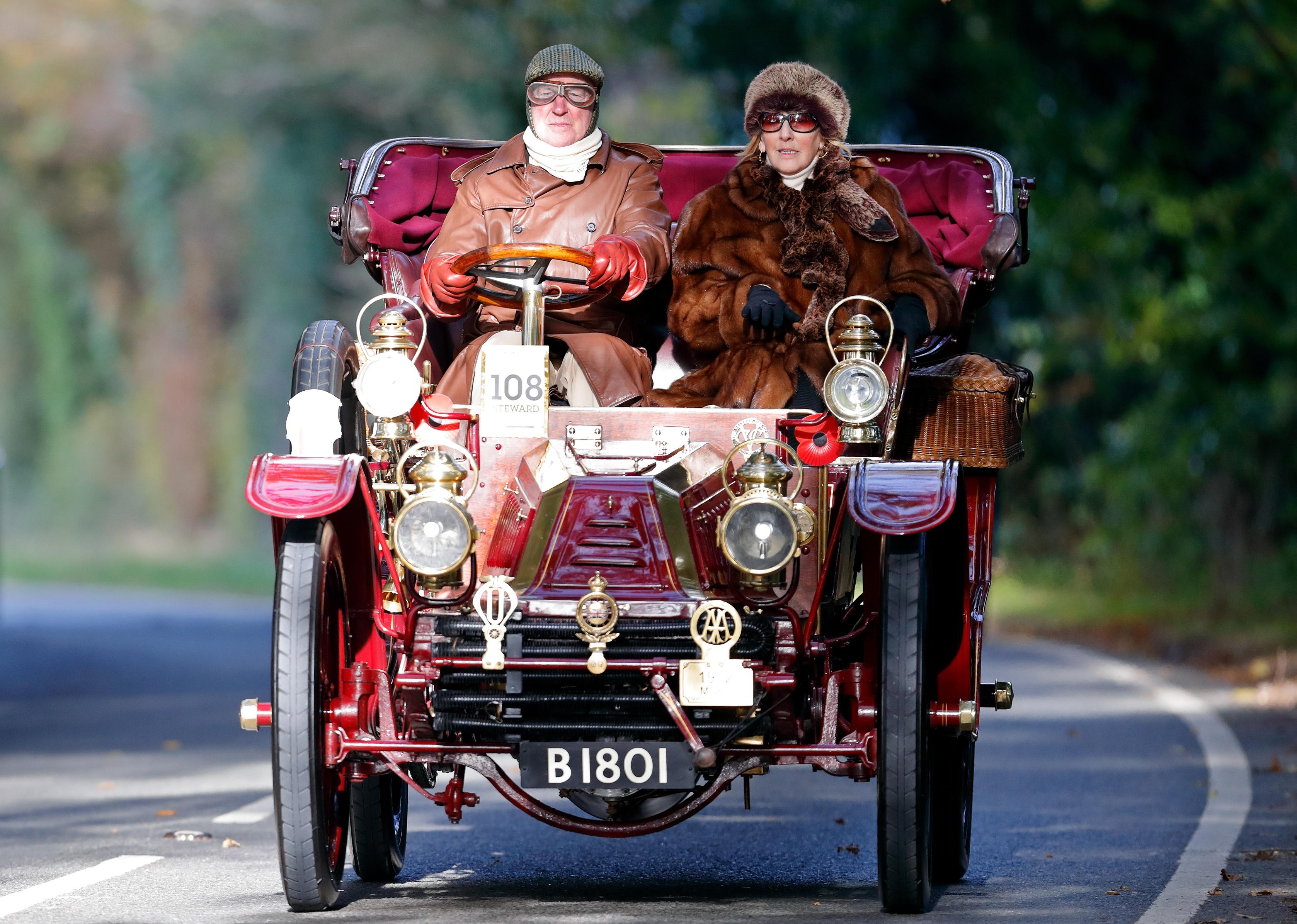 A man in a leather jacket and goggles and a woman in a fur coat driving a 1902 Mors classic car.