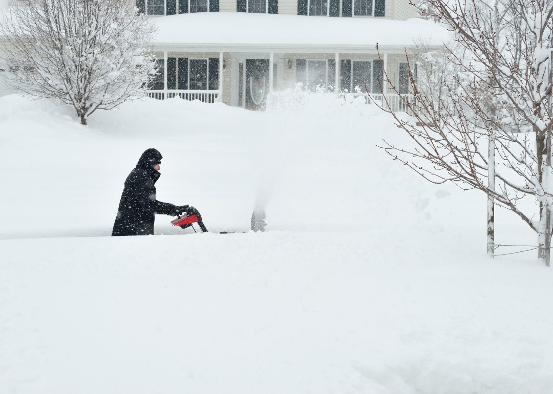 A figure uses a snowblower in deep snow outside a home.