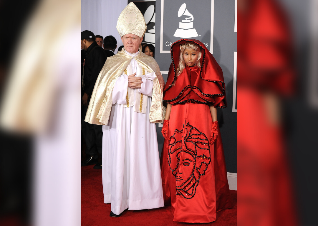Nicki Minaj in a red silk gown and hood with black bead details posing with a Pope lookalike.