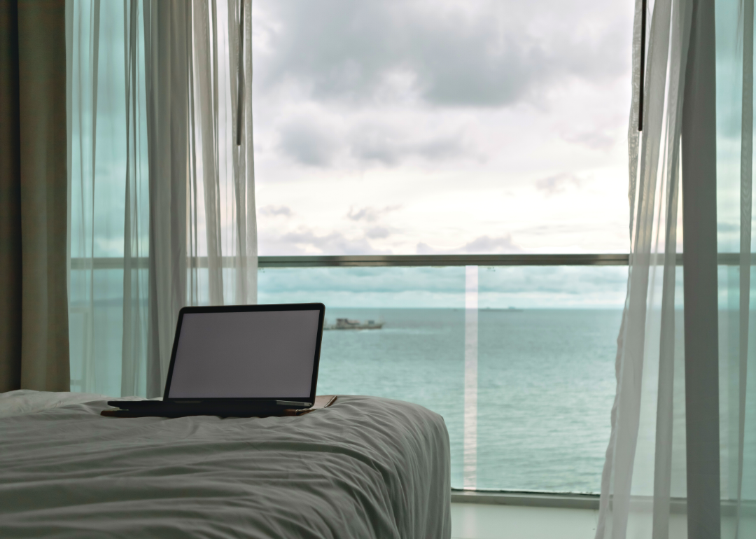 A laptop lies unattended on a hotel bed with a view of the ocean in the background.