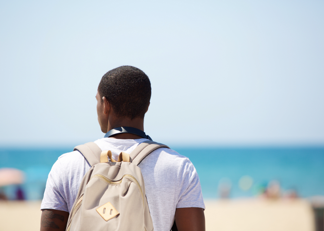 A man wearing a backpack looks out at the beach and sea.