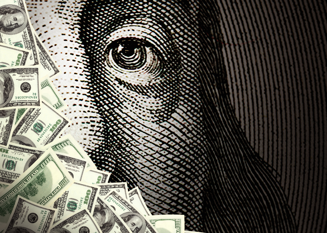 A pile of money sits a top a close up view of Benjamin Franklin as seen on a $100 bill. 
