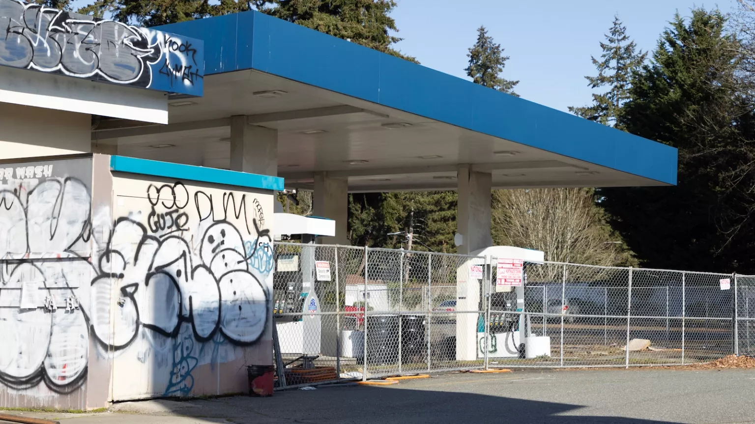 A gas station is surrounded by wire fencing and has graffiti on its walls.