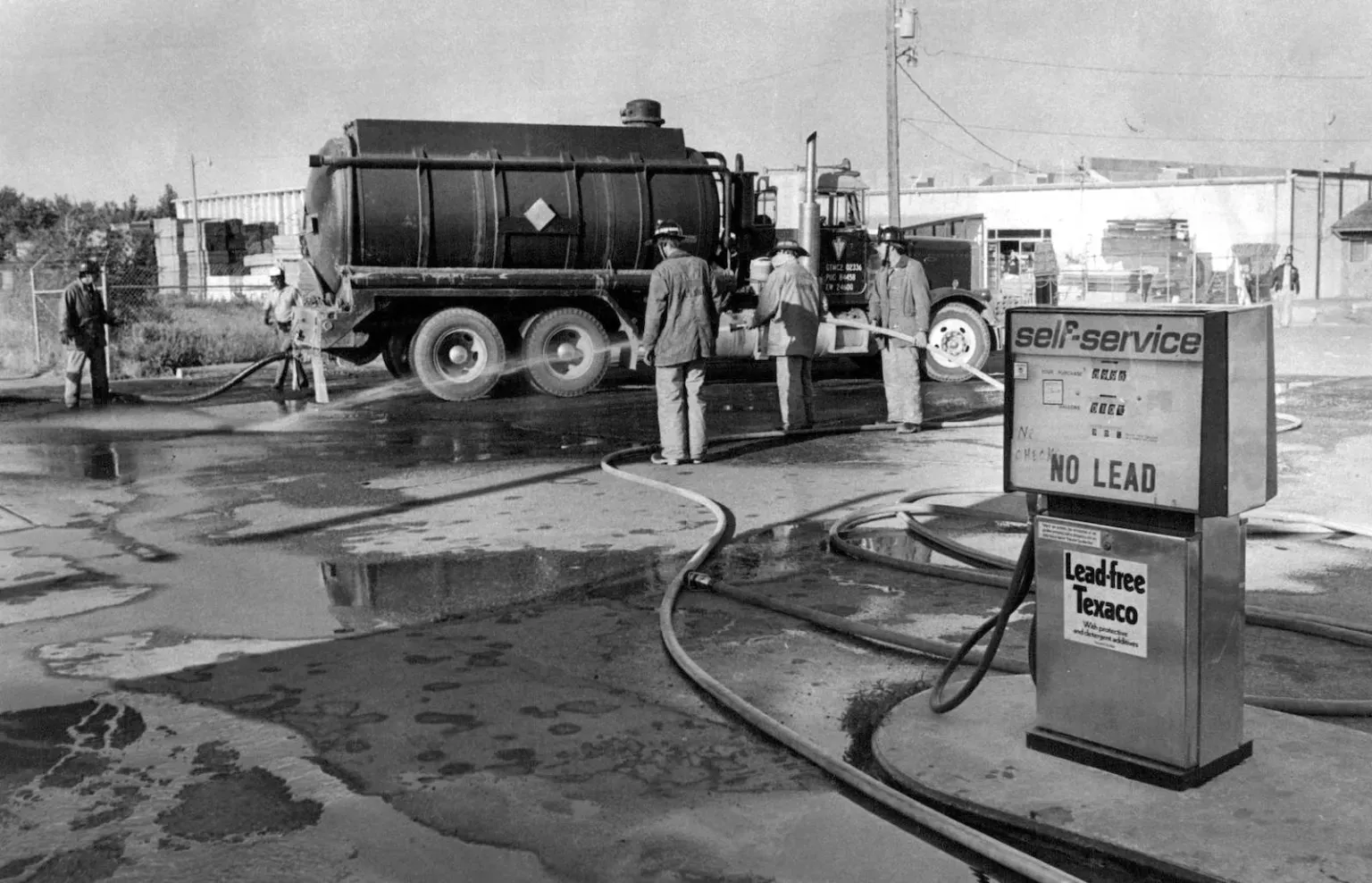Firefighters spray liquid on a paved area near a large truck, with a gas pump in the foreground.