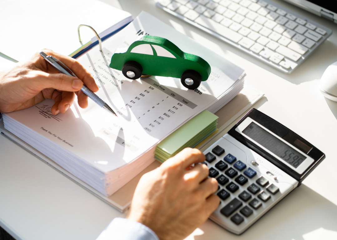 A desktop on which a person is working through financial documents with a pen and calculator; a small green car rests atop the paperwork.