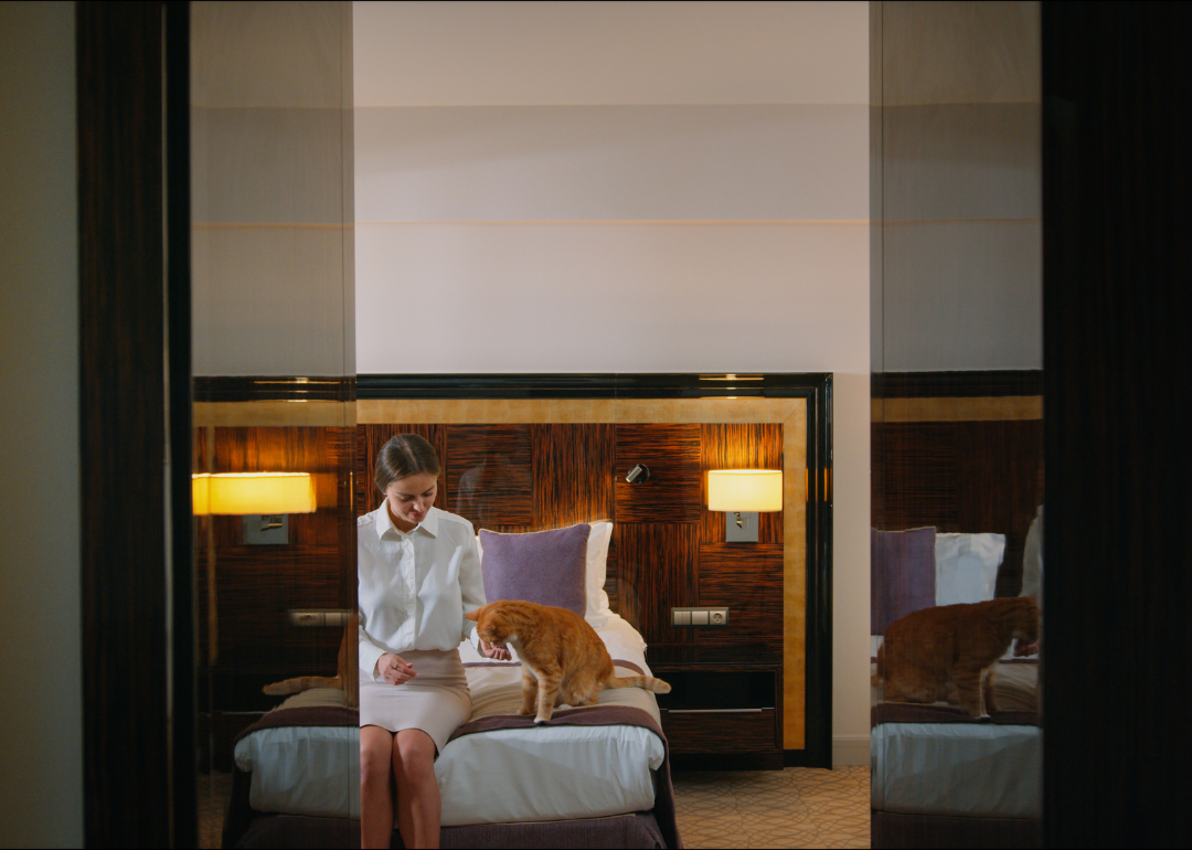 A woman and ginger cat sit on the bed in a hotel room.