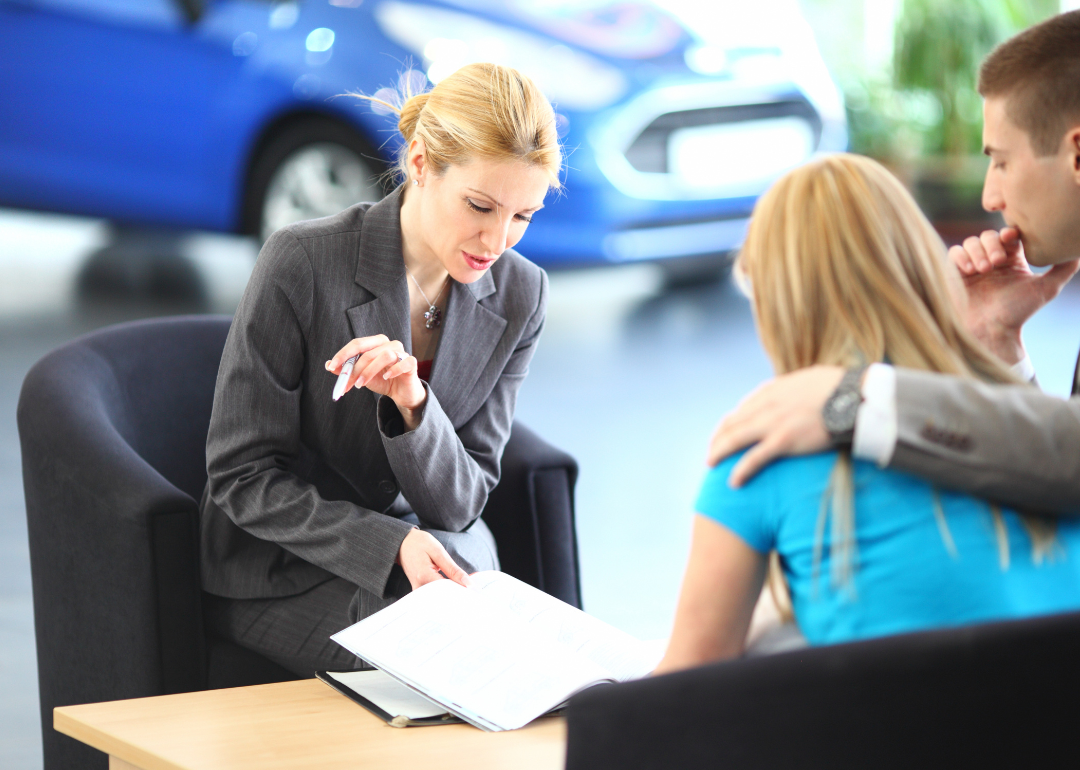 A loan officer explaining some paperwork to a young couple seated at a desk in a car dealership.